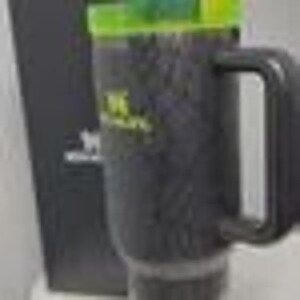 May include: A black travel mug with a green and yellow lid and handle. The mug has a textured surface and a logo on the side. The mug is next to a black box.