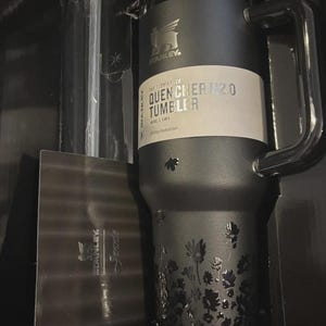 Puede incluir: Un vaso Stanley Quencher H2.0 negro con asa, con un dise&ntilde;o floral y el texto "THE FLOWSTATE QUENCHER H2.0 TUMBLER". El vaso est&aacute; empaquetado en una caja negra.