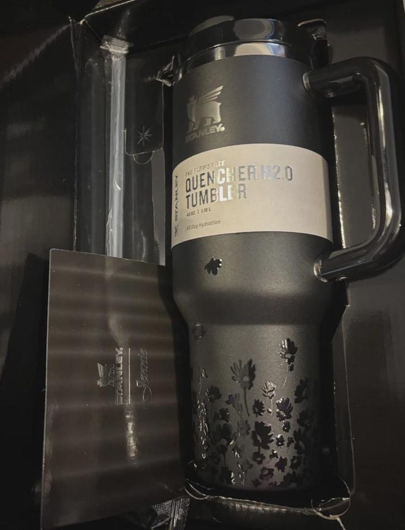 Puede incluir: Un vaso Stanley Quencher H2.0 negro con asa, con un dise&ntilde;o floral y el texto "THE FLOWSTATE QUENCHER H2.0 TUMBLER". El vaso est&aacute; empaquetado en una caja negra.