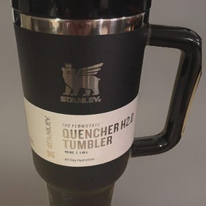 Puede incluir: Vaso Stanley Quencher H2.0 negro con un asa grande. El vaso tiene un borde plateado y una etiqueta blanca con el texto "THE FLOWSTATE QUENCHER H2.0 TUMBLER 1,18 L All Day Hydration". El logotipo de Stanley es visible.
