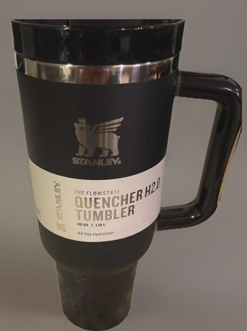 Puede incluir: Vaso Stanley Quencher H2.0 negro con un asa grande. El vaso tiene un borde plateado y una etiqueta blanca con el texto "THE FLOWSTATE QUENCHER H2.0 TUMBLER 1,18 L All Day Hydration". El logotipo de Stanley es visible.