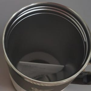Puede incluir: Una taza aislada negra y plateada con asa. El interior es negro, y el borde y el asa son plateados. La taza tiene una banda blanca alrededor de la base con el nombre y el logotipo de la marca. La taza est&aacute; dise&ntilde;ada para mantener las bebidas calientes o fr&iacute;as.