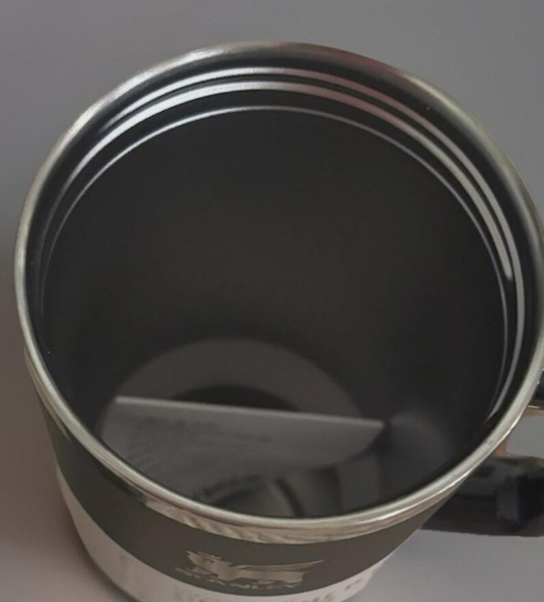 Puede incluir: Una taza aislada negra y plateada con asa. El interior es negro, y el borde y el asa son plateados. La taza tiene una banda blanca alrededor de la base con el nombre y el logotipo de la marca. La taza est&aacute; dise&ntilde;ada para mantener las bebidas calientes o fr&iacute;as.