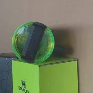 May include: A clear Stanley tumbler lid with a black handle and a neon green rim rests on a bright green box. The Stanley logo is visible on the box. The lid is partially transparent, showing the tumbler's interior.