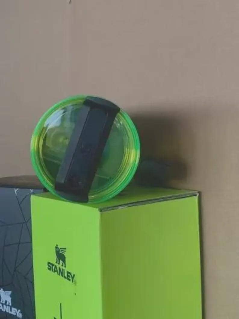 May include: A clear Stanley tumbler lid with a black handle and a neon green rim rests on a bright green box. The Stanley logo is visible on the box. The lid is partially transparent, showing the tumbler's interior.