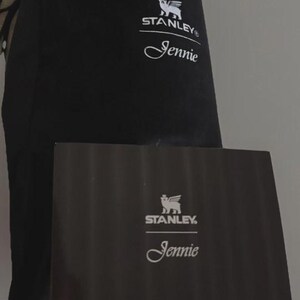 Puede incluir: Juego de bolsa y caja negros de la marca Stanley. La bolsa y la caja presentan el logotipo de Stanley y el nombre "Jennie" en escritura blanca. La caja es rectangular y parece ser de cart&oacute;n.