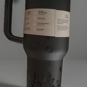 Puede incluir: Taza de viaje negra con asa, detalles florales y los nombres "Jennie" y "Ruby". La taza tiene tapa y una etiqueta de papel con informaci&oacute;n del producto. Dise&ntilde;ada para bebidas fr&iacute;as y calientes.