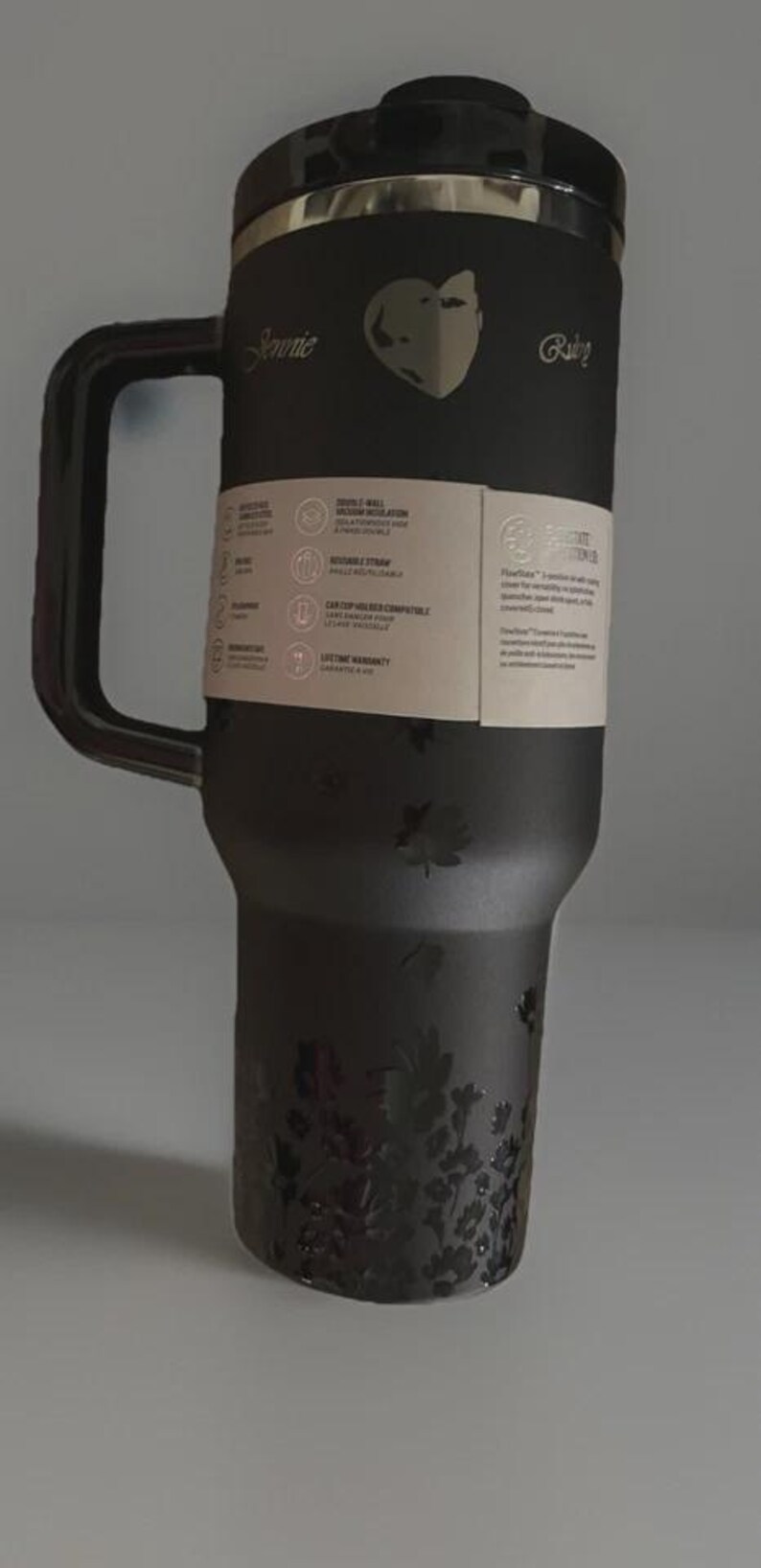 Puede incluir: Taza de viaje negra con asa, detalles florales y los nombres "Jennie" y "Ruby". La taza tiene tapa y una etiqueta de papel con informaci&oacute;n del producto. Dise&ntilde;ada para bebidas fr&iacute;as y calientes.