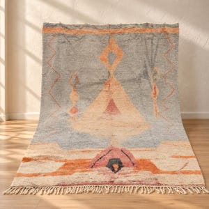 May include: A rectangular area rug featuring a gray background with a large central peach-colored diamond and triangle motif. The design includes orange zigzag lines, a dark pink base element, and a fringe border along the bottom edge.