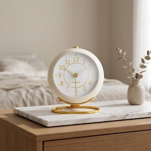 May include: Cream-colored, round analog alarm clock with gold-colored accents. The clock sits on a marble surface atop a wooden nightstand. The clock face has gold numerals and hands. A small vase with dried stems is to the right of the clock.