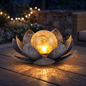 May include: A decorative outdoor solar light shaped like a lotus flower. The flower has gray petals surrounding a glowing, textured, amber-colored glass orb. The light sits on a wooden table, with a blurred background of outdoor lights and foliage.