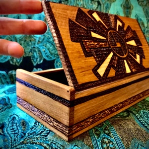 May include: A wooden box with a hinged lid. The lid is open and features a carved design of a sun with rays. The box is made of light brown wood with dark brown accents.