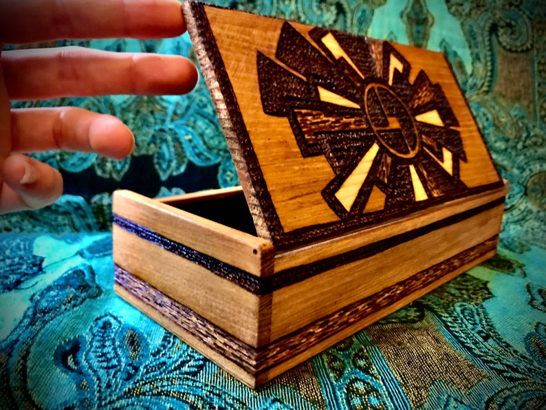 May include: A wooden box with a hinged lid. The lid is open and features a carved design of a sun with rays. The box is made of light brown wood with dark brown accents.