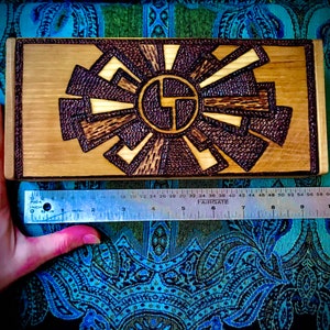 May include: A wooden box with a geometric design burned into the surface. The design is in shades of brown and black, and it resembles a sunburst.