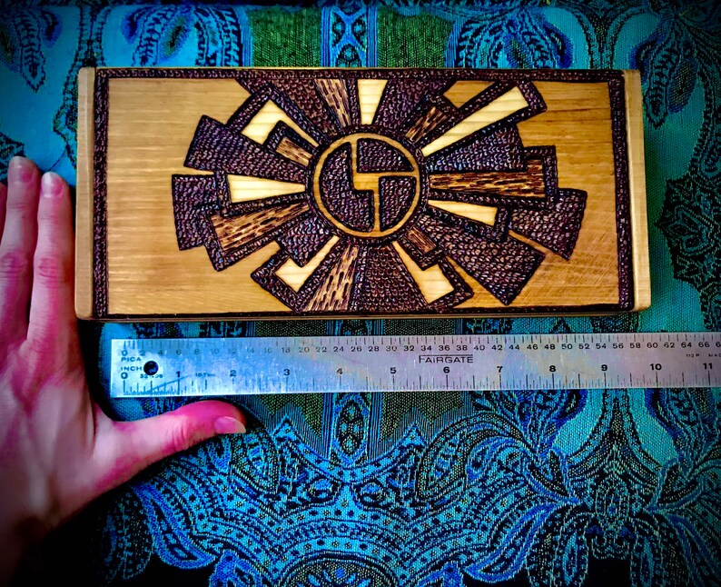 May include: A wooden box with a geometric design burned into the surface. The design is in shades of brown and black, and it resembles a sunburst.