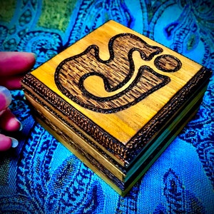May include: A small wooden box with a dark brown trim and a carved design of a stylized symbol on the top. The box is made of light wood and has a natural finish.