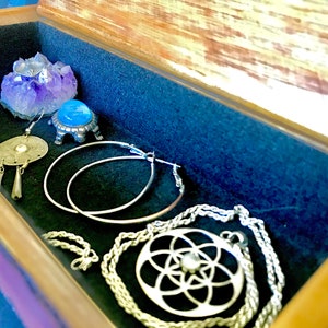 May include: A wooden box with a velvet lining, containing a variety of silver jewelry, including a necklace with a geometric pendant, hoop earrings, and dangle earrings. A small amethyst crystal sits on a silver stand.