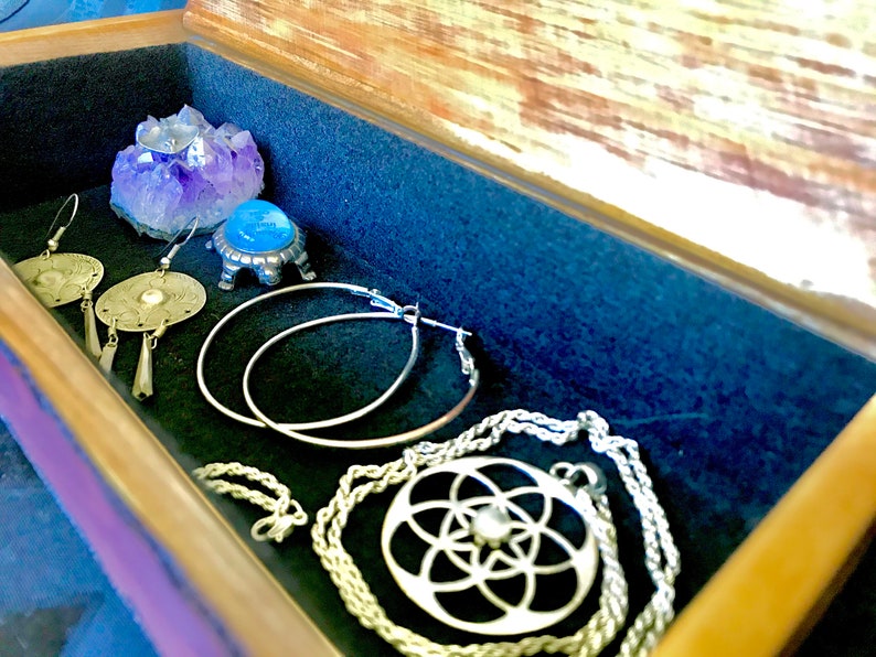 May include: A wooden box with a velvet lining, containing a variety of silver jewelry, including a necklace with a geometric pendant, hoop earrings, and dangle earrings. A small amethyst crystal sits on a silver stand.