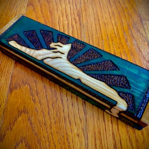 May include: A rectangular wooden box with a detailed carving of a figure reaching upwards, set against a sunburst design. The box features a green and brown color scheme, with a natural wood finish.