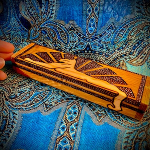 May include: A rectangular wooden box with a carved design of a figure reaching upwards, set against a sunburst pattern. The box is light brown with darker brown accents and is displayed on a blue patterned fabric.
