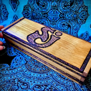 May include: A wooden box with a purple Om symbol inlay. The box has a dark wood finish and a light wood trim. The box is sitting on a blue and black patterned fabric.