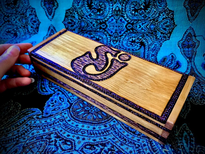 May include: A wooden box with a purple Om symbol inlay. The box has a dark wood finish and a light wood trim. The box is sitting on a blue and black patterned fabric.
