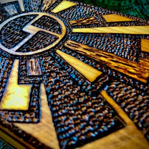 May include: A wooden box with a geometric design carved into the top. The design is in shades of brown and black, and features a circle with a stylised letter "L" inside.