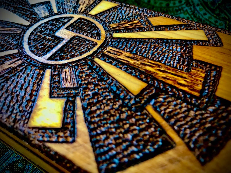 May include: A wooden box with a geometric design carved into the top. The design is in shades of brown and black, and features a circle with a stylised letter "L" inside.