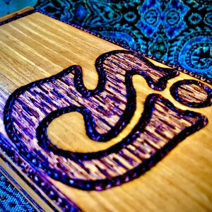 May include: A wooden box with a purple Om symbol carved into the top. The box is made of light wood and has a dark brown stain.