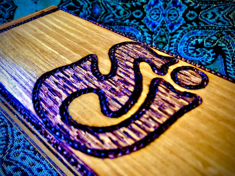 May include: A wooden box with a purple Om symbol carved into the top. The box is made of light wood and has a dark brown stain.