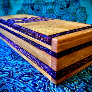 May include: A wooden box with purple and brown inlay. The box has a rectangular shape and is made of light wood. The box is decorated with purple and brown inlay.