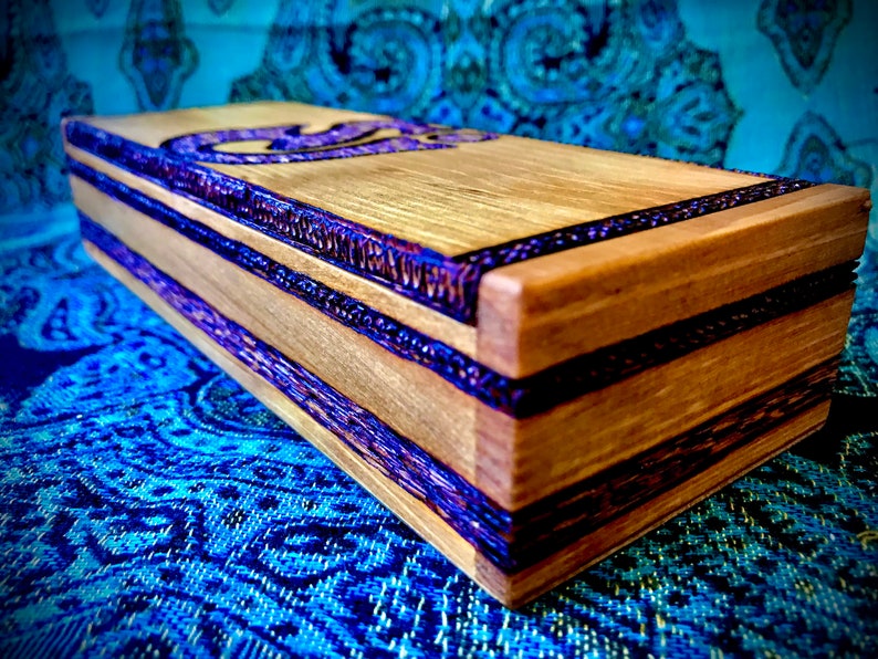 May include: A wooden box with purple and brown inlay. The box has a rectangular shape and is made of light wood. The box is decorated with purple and brown inlay.