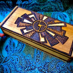 May include: A wooden box with a decorative, intricate design burned into the top. The design features a circular pattern with radiating lines and a central symbol.