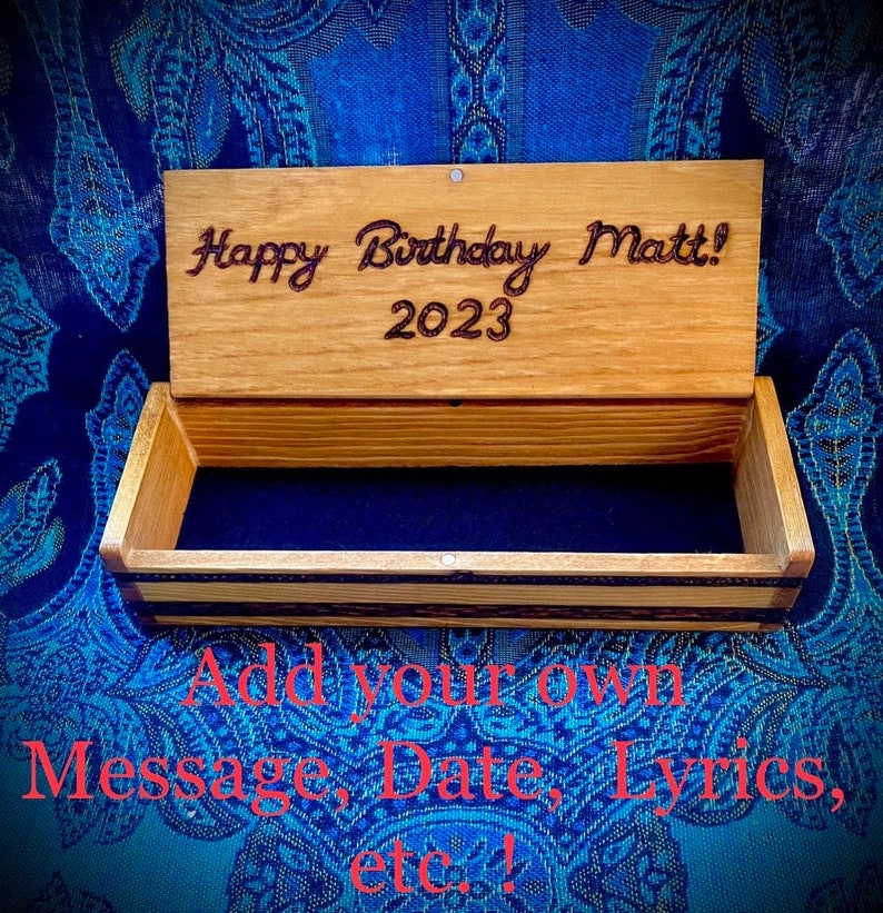 May include: A wooden box with a hinged lid. The lid is open and has the text "Happy Birthday Matt! 2023" engraved on it. The box is empty and has a dark blue velvet lining.