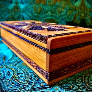 May include: A wooden box with a dark brown wood inlay design. The box has a rectangular shape and is made of light brown wood. The box is sitting on a blue and green patterned fabric.