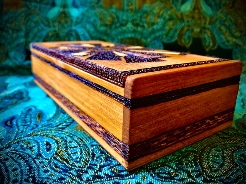 May include: A wooden box with a dark brown wood inlay design. The box has a rectangular shape and is made of light brown wood. The box is sitting on a blue and green patterned fabric.