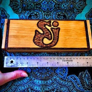 May include: A wooden box with a dark brown wood inlay around the edges. The box has a woodburned design of the letters "SJ" in a stylized font. The box is approximately 6 inches long.