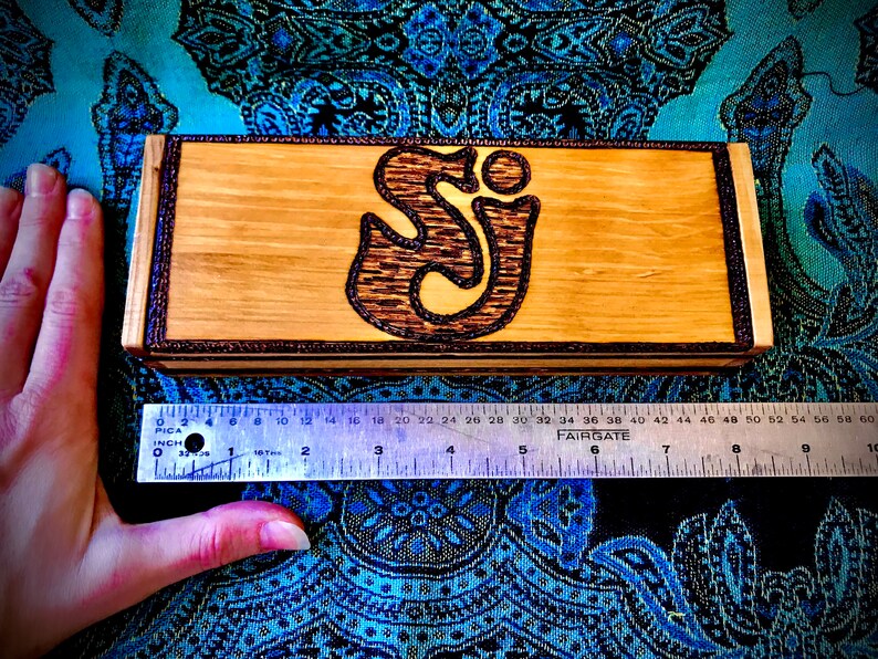 May include: A wooden box with a dark brown wood inlay around the edges. The box has a woodburned design of the letters "SJ" in a stylized font. The box is approximately 6 inches long.