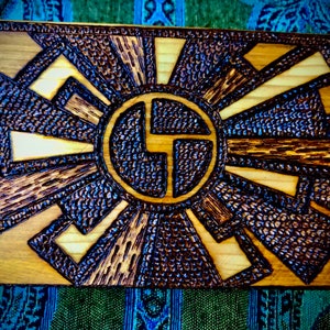 May include: A wooden wall art piece with a dark brown woodburned design of a stylised sun with rays. The sun has a circle in the centre with a stylised letter "G" inside.