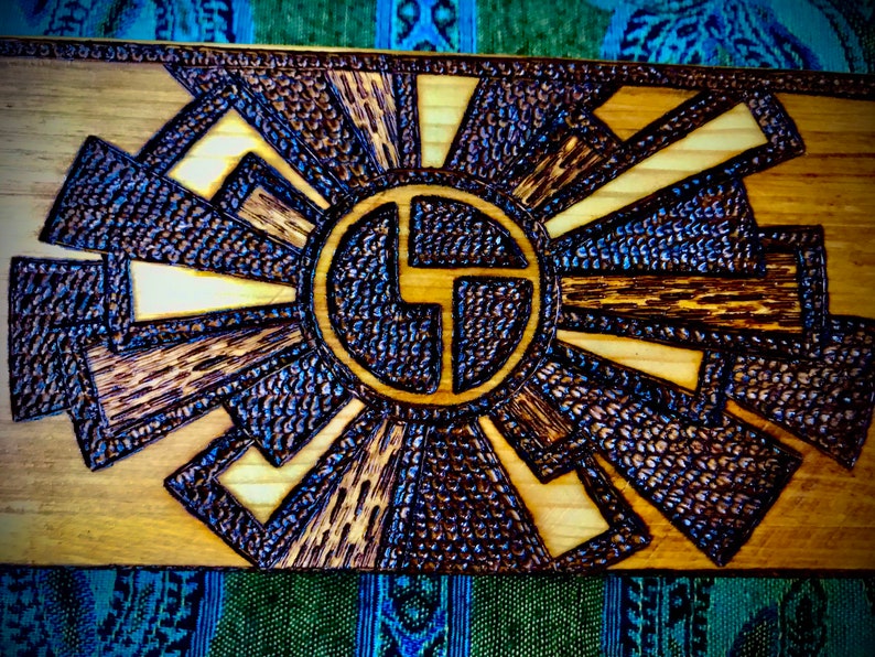 May include: A wooden wall art piece with a dark brown woodburned design of a stylised sun with rays. The sun has a circle in the centre with a stylised letter "G" inside.