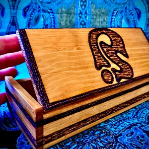 May include: A wooden box with a dark brown wood inlay and a wood-burned design of a stylized ear on the lid. The box is open and the inside is visible.