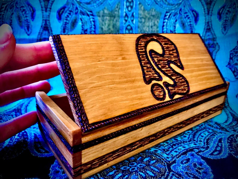 May include: A wooden box with a dark brown wood inlay and a wood-burned design of a stylized ear on the lid. The box is open and the inside is visible.