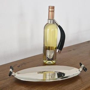 Puede incluir: Un soporte plateado para botellas de vino con un asa negra y una bandeja ovalada a juego. La botella contiene un líquido amarillo claro. La bandeja tiene detalles negros en las asas. Los artículos están sobre una superficie de madera.