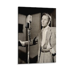 May include: Black and white photograph of a man in a suit standing in front of a microphone. The man is wearing a patterned tie and a light-colored shirt. The microphone is a vintage style, and the background is a recording studio.