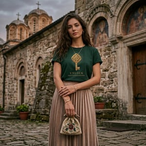 May include: A woman wearing a dark green t-shirt with a gold key graphic and the text "UNLOCK YOUR UNLIMITED POTENTIAL". She is also wearing a gold necklace with a key pendant, a pleated skirt, and holding a small embroidered bag.