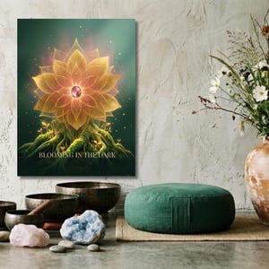 Puede incluir: Una impresión en lienzo con un diseño de flor de loto dorada con un centro rojo, enraizada en una base musgosa. El texto "BLOOMING IN THE DARK" está en la parte inferior. La impresión se muestra cerca de un cojín de meditación verde y otros elementos espirituales.
