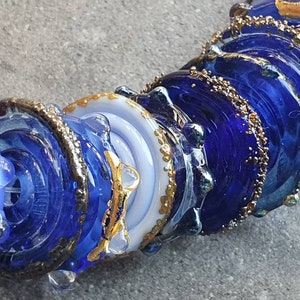 Op de afbeelding: Close-up van een blauwe en witte glazen kralen met gouden accenten. De kraal maakt deel uit van een grotere kralenstreng, en de kleuren zijn levendig en opvallend.