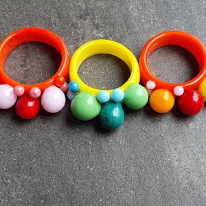 May include: Three colorful rings with a beaded design. Each ring is made of a different color: orange, yellow, and green. The beads are in various colors, including pink, red, orange, yellow, green, and blue.