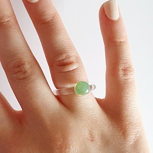 Puede incluir: Un anillo transparente con una piedra de vidrio verde claro. El anillo está en un dedo.