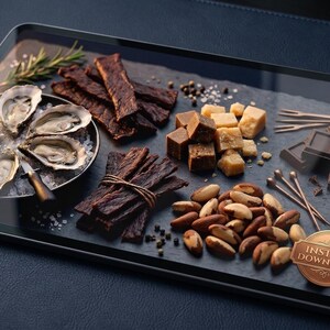 May include: A gourmet food platter featuring oysters on ice, jerky, cheese cubes, chocolate squares, and Brazil nuts. Garnished with rosemary and sea salt, the arrangement is presented on a dark slate tray.